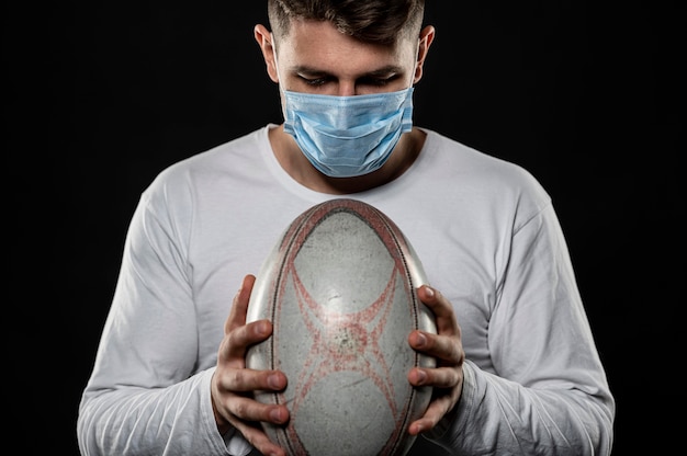 Kostenloses Foto männlicher rugbyspieler, der ball hält, während er medizinische maske trägt