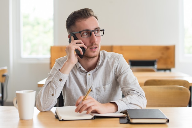 Männlicher Student, der am Telefon spricht und Hausarbeit am Schreibtisch tut