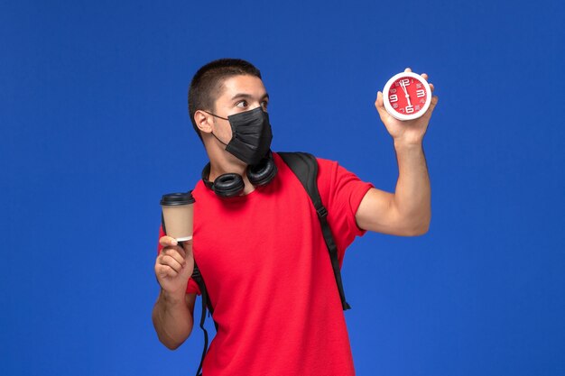 Männlicher Student der Vorderansicht im roten T-Shirt, das Maske mit Rucksack hält, der Uhrenkaffee auf blauem Schreibtisch hält.