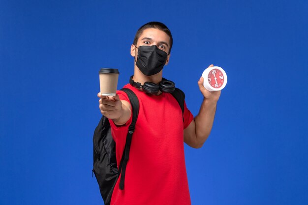 Männlicher Student der Vorderansicht im roten T-Shirt, das Maske mit Rucksack hält, der Uhrenkaffee auf hellblauem Hintergrund hält.