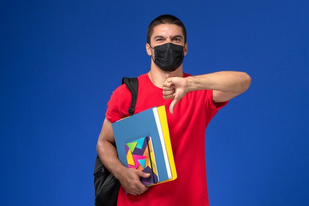 Männlicher Student der Vorderansicht im roten T-Shirt, das Rucksack in der schwarzen sterilen Maske hält, die Copybooks hält, zeigt Zeichen anders als Zeichen auf blauem Hintergrund.