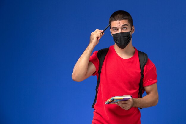 Männlicher Student der Vorderansicht im roten T-Shirt, das Rucksack in der schwarzen sterilen Maske hält, die das Heft und das Stiftdenken auf dem blauen Hintergrund hält.