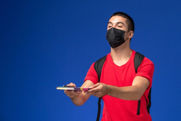 Männlicher Student der Vorderansicht im roten T-Shirt, das Rucksack in der schwarzen sterilen Maske hält, die Stift und Heft auf dem blauen Schreibtisch hält.