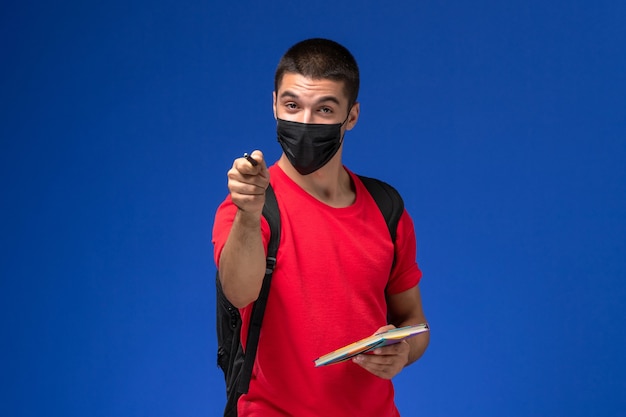 Männlicher Student der Vorderansicht im roten T-Shirt, das Rucksack in schwarzer steriler Maske trägt, die Stift und Heft auf dem blauen Hintergrund hält.