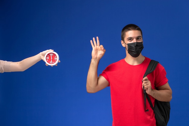 Männlicher Student der Vorderansicht im roten T-Shirt, das Rucksack mit Maske trägt, die auf blauem Hintergrund aufwirft.