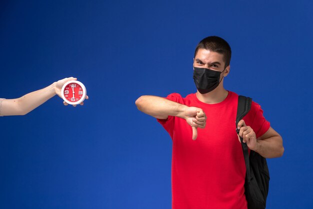 Männlicher Student der Vorderansicht im roten T-Shirt, das Rucksack mit Maske trägt, die auf blauem Hintergrund aufwirft.