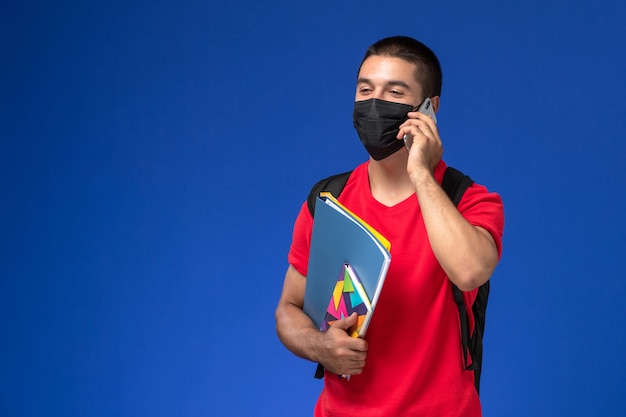 Männlicher Student der Vorderansicht im roten T-Shirt, der Rucksack mit Maske hält, die Copybook-Dateien hält, die auf dem Telefon auf dem blauen Hintergrund sprechen.
