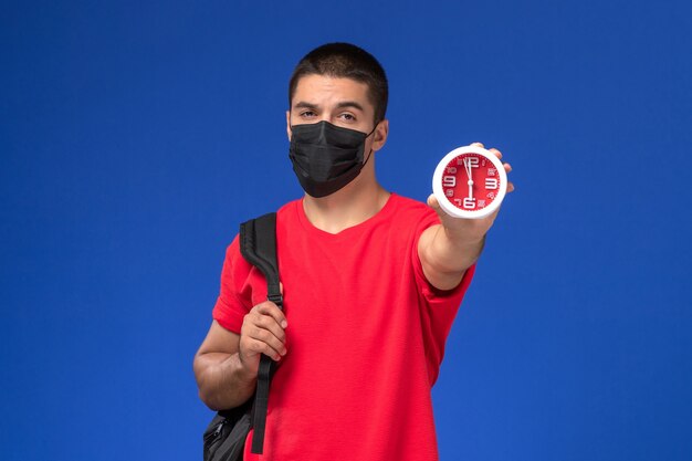 Männlicher Student der Vorderansicht im roten T-Shirt, der Rucksack mit Maske hält Uhren auf dem blauen Hintergrund trägt.