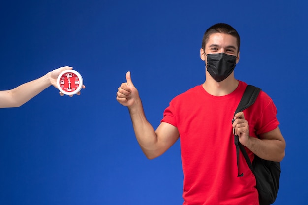 Männlicher Student der Vorderansicht im roten T-Shirt, der Rucksack mit Maske trägt, die auf dem blauen Schreibtisch aufwirft.