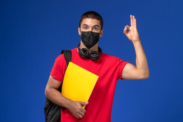 Männlicher Student der Vorderansicht in der roten T-Shirt tragenden Maske mit dem Rucksack, der gelbe Datei auf dem blauen Hintergrund hält.