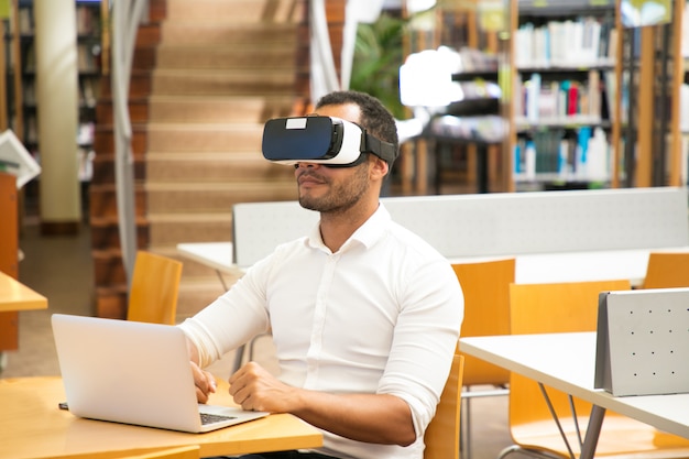 Männlicher Student, der VR-Kopfhörer während der Arbeit in der Bibliothek verwendet