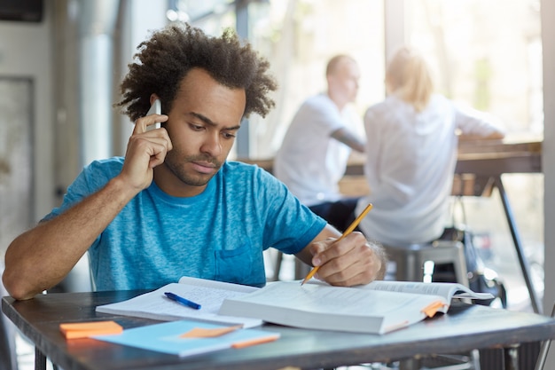 Männlicher Student mit afrikanischer Frisur, der am hölzernen Schreibtisch sitzt und mit seinem besten Freund am Smartphone spricht, die neuesten Nachrichten bespricht und ernsthaft im Lehrbuch schaut, unterstreicht etwas mit Bleistift