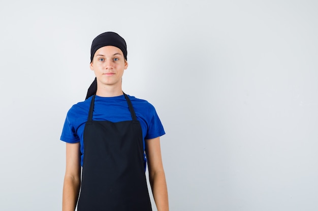 Männlicher Teenagerkoch im T-Shirt, Schürze posiert im Stehen und sieht selbstbewusst aus, Vorderansicht.