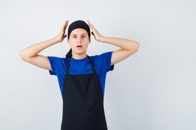 Männlicher Teenagerkoch mit den Händen in der Nähe des Kopfes, öffnet den Mund in T-Shirt, Schürze und sieht entsetzt aus, Vorderansicht.