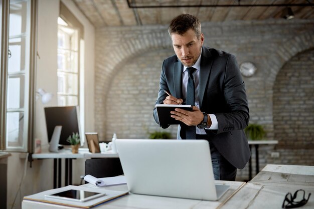 Männlicher Unternehmer, der eine E-Mail auf einem Computer liest und sich Notizen macht, während er im Büro arbeitet
