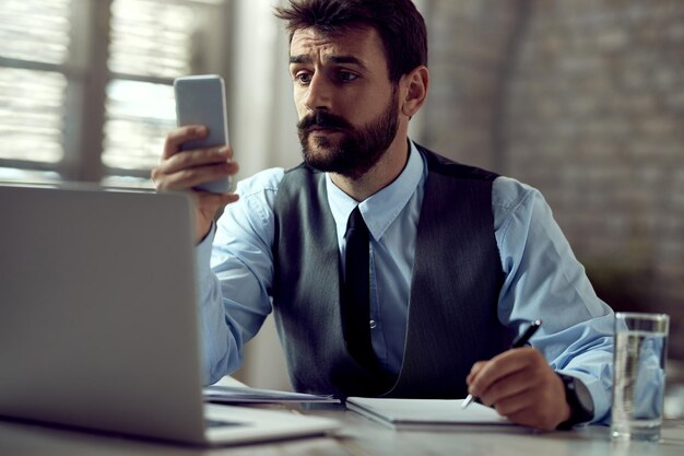 Männlicher Unternehmer, der Handy und Textnachrichten verwendet, während er sich Notizen im Büro macht