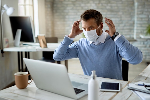 Männlicher Unternehmer, der im Büro arbeitet und eine schützende Gesichtsmaske aufsetzt