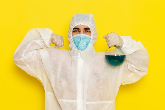 Männlicher wissenschaftlicher Arbeiter der Vorderansicht im speziellen Schutzanzug, der Flasche mit blauer Lösung hält, die auf gelber Oberfläche biegt