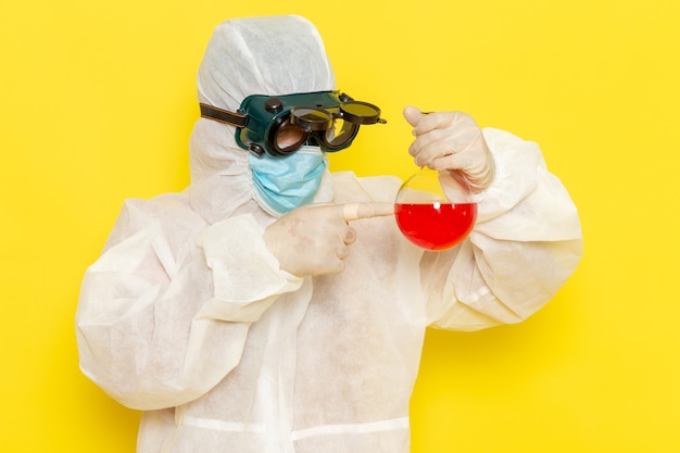 Männlicher wissenschaftlicher Arbeiter der Vorderansicht im speziellen Schutzanzug, der Flasche mit roter Lösung auf gelber Oberfläche hält