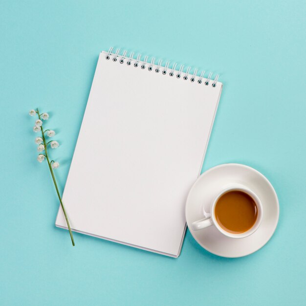 Maiglöckchenblumenzweig auf weißem gewundenem Notizblock mit Kaffeetasse auf blauem Hintergrund