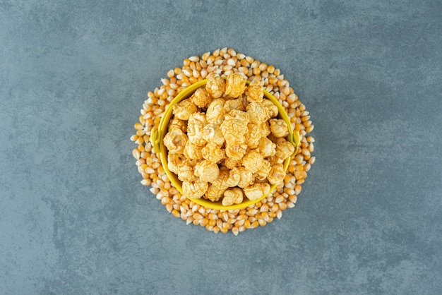 Maiskörner um einen Eimer mit Popcorn-Süßigkeiten, die mit Karamell auf Marmorhintergrund beschichtet sind. Foto in hoher Qualität