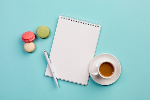 Makronen mit gewundenem Notizblock, Stift und Kaffeetasse auf blauem Hintergrund