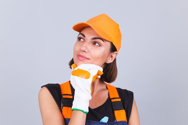 Malerarbeiterfrau in Overalls und Handschuhen auf grauer Wand glückliches positives Lächeln nachdenklicher Blick beiseite