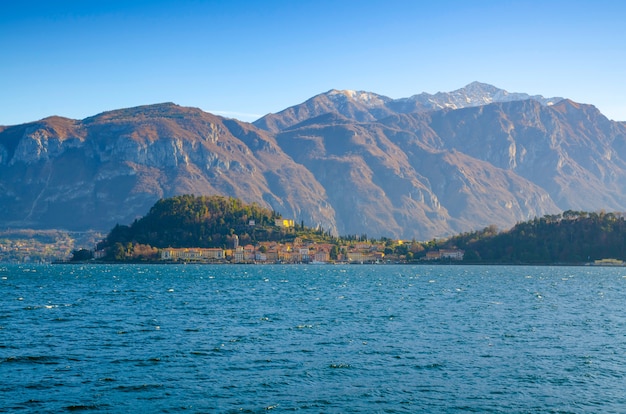 Malerischer See mit einem Küstendorf am Horizont und den Bergen gegen einen blauen Himmel