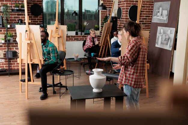 Malerlehrer zeigt Modell für Zeichenkurs und erklärt Illustrationstechnik während des Kunstunterrichts im Kreativitätsstudio. Multiethnisches Team skizziert Vase auf Malleinwand mit grafischem Bleistift