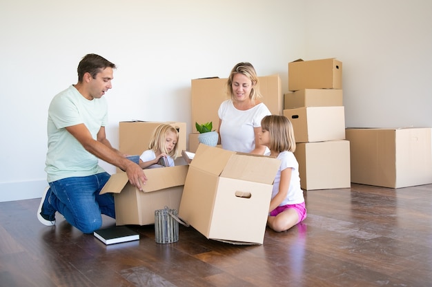 Mama, Papa und kleine Töchter packen Dinge in einer neuen Wohnung aus, sitzen auf dem Boden und nehmen Gegenstände aus offenen Kisten