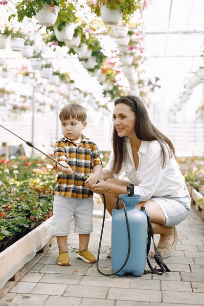 Mama und ihr Sohn mit einem blauen Wassersprüher im Gewächshaus. Kleiner Kleinkindjunge, der Blumen in einem Gewächshaus gießt