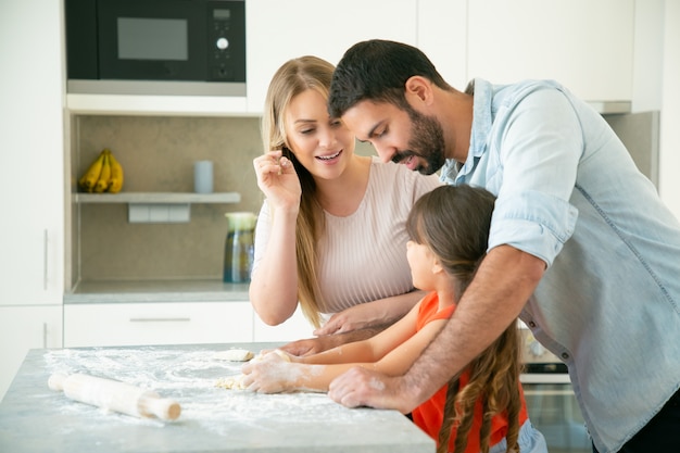 Mama und Papa lehren Kind, Teig auf Küchentisch mit Mehl unordentlich zu kneten. Junges Paar und ihr Mädchen backen Brötchen oder Kuchen zusammen. Familienkochkonzept