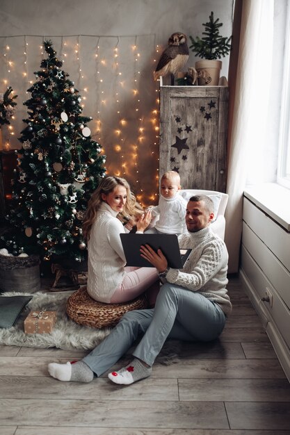 Mama und Papa sehen Cartoons auf Laptop mit ihrem Baby in der Nähe des Weihnachtsbaumes