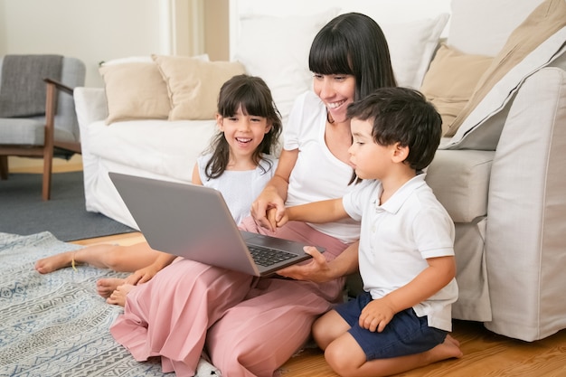 Mama und zwei Kinder schauen sich einen lustigen Film an, während sie im Wohnzimmer auf dem Boden sitzen, einen Laptop benutzen und lachen.