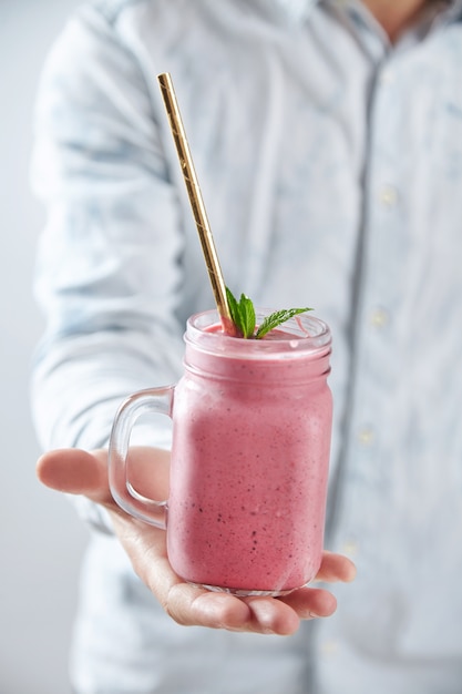 Man Hand bietet ein rustikales Glas mit kaltem, frischem Beeren-Smoothie mit goldenem Stroh und Minzblättern an. Sommer Erfrischungsgetränk Nah fokussiert, isoliert