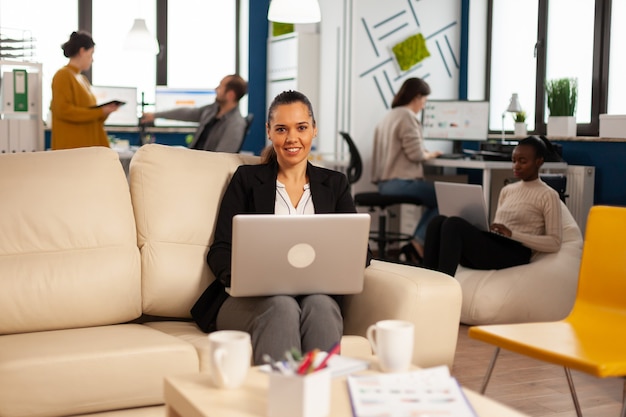 Managerin schreibt auf Laptop und schaut lächelnd in die Kamera, während verschiedene Kollegen im Hintergrund arbeiten working