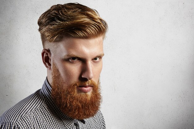 Manful europäischer Hipster im karierten Hemd, das ernst schaut. Sein stilvoller Haarschnitt und sein gut geschnittener blonder Bart sagen, dass er ein treuer Kunde des Friseursalons ist und sich um sein Aussehen kümmert.
