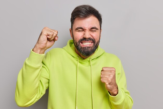 Mann ballt die Faust vor Glück freut sich, dass sich etwas anfühlt, als würde der Gewinner die Zähne zusammenbeißen, gekleidet in einem lässigen Hoodie isoliert auf Grau