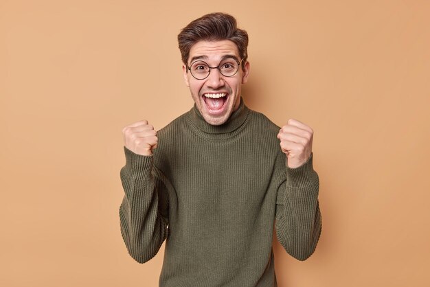 Mann ballt Fäuste Triumphe von erfolgreichen Deals oder großartigen Neuigkeiten ist freudig und begeistert trägt einen lässigen Pullover und eine Brille posiert gegen Beige fängt den Traum ein