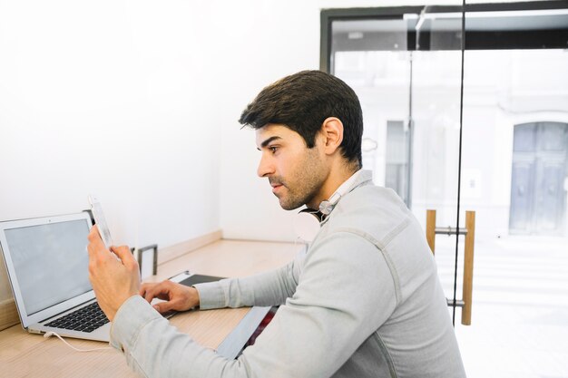 Mann, der am Laptop unter Verwendung des Smartphone sitzt