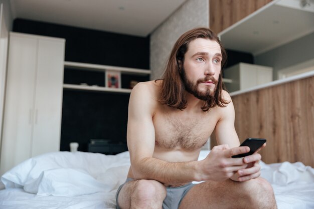 Mann, der auf Bett sitzt und Handy am Morgen benutzt