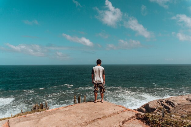 Mann, der auf den Felsen steht, die durch Grün und das Meer unter Sonnenlicht und einem blauen Himmel umgeben sind