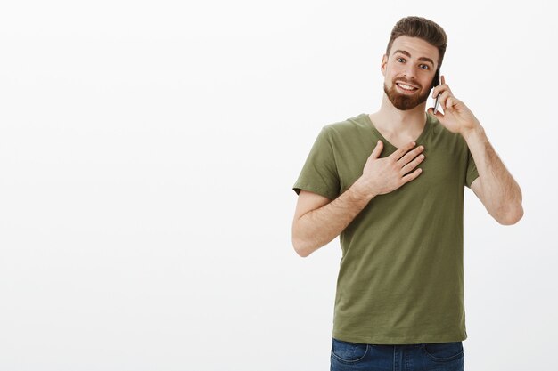Mann, der dankbar und erfreut empfängt, Telefonanruf zu erhalten, der auf Smartphone spricht, das mit bday über Handy beglückwünscht wird, das Hand auf Brust dankbar hält und glücklich über weiße Wand lächelt