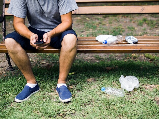 Mann, der das Mobiltelefon sitzt auf Bank nahe Plastikabfall am Park verwendet