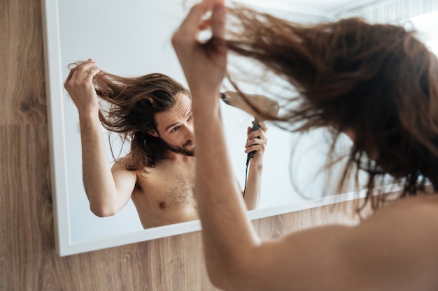 Mann, der den Spiegel betrachtet und Haare mit Haartrockner trocknet