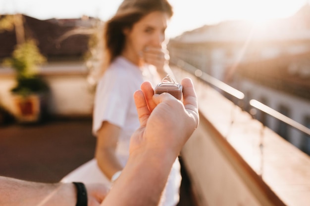 Mann, der Ehering vor erstauntem glücklichem Mädchen hält, das den Mund mit der Hand bedeckt. Romantisches Foto einer charmanten Frau, die am frühen Abend beim Date mit ihrem Freund im Jubiläum auf dem Dach steht.