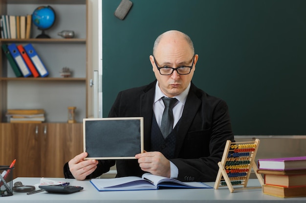 Mann, der eine Brille trägt und an der Schulbank vor der Tafel im Klassenzimmer sitzt und eine Tafel zeigt, die die Lektion erklärt, die Lektion mit ernstem Gesicht erklärt