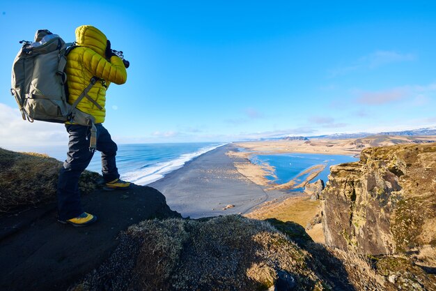 Mann, der eine gelbe Jacke trägt, die auf einem Felsen steht, während ein Foto der schönen Landschaft macht