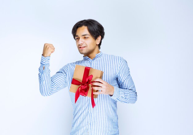 Mann, der eine Geschenkbox aus Karton mit rotem Band hält und seine Faust als erfolgreicher Mensch zeigt. Foto in hoher Qualität