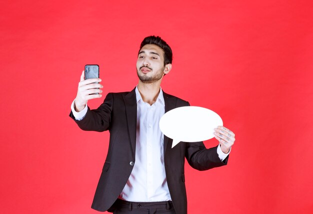 Mann, der eine leere ovale Infotafel hält und mit dem Telefon spricht oder einen Videoanruf tätigt.
